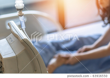 Saline intravenous (IV) drip for medical treatment and nursing patient in hospital with blurred family caregiver or nurse caregiving ill patient on bed in ward 63916550