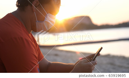 COVID -19. Man wearing mask on sits on the beach uses mobile phone at sunset time. Coronavirus epidemic COVID -19. Man wearing mask on sits on the beach uses mobile phone at sunset time. Coronavirus epidemic 63919695