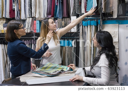 Female seller talking with buyer in store of fabrics and accessories for curtains, interior showroom. Woman looking fabrics for curtains upholstery Female seller talking with buyer in store of fabrics and accessories for curtains, interior showroom. Woman looking fabrics for curtains upholstery 63923113