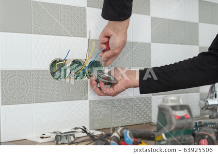 Renovation and construction in kitchen, close-up of electricians hand installing outlet on wall with ceramic tiles using professional tools 63925506