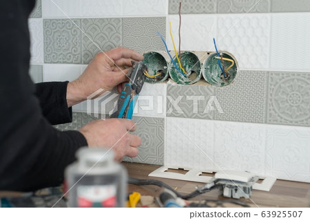 Renovation and construction in kitchen, close-up of electricians hand installing outlet on wall with ceramic tiles using professional tools 63925507