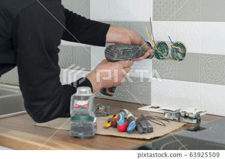 Renovation and construction in kitchen, close-up of electricians hand installing outlet on wall with ceramic tiles using professional tools 63925509