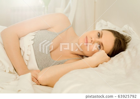 Young beautiful woman lying in bed on pillow looking in camera smiling, bright sunny bedroom interior 63925792