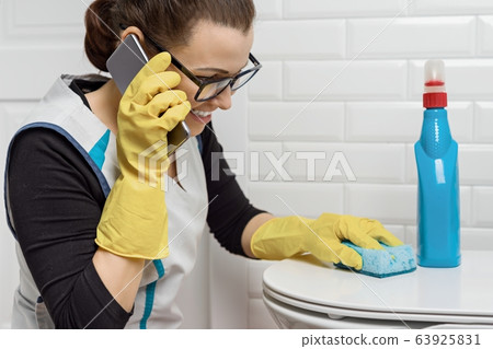 Adult positive woman doing cleaning with detergents. Woman in glasses, professional uniform in bathroom, toilets, woman laughing and talking on phone Adult positive woman doing cleaning with detergents. Woman in glasses, professional uniform in bathroom, toilets, woman laughing and talking on phone 63925831