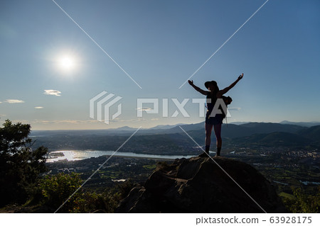 Silhouette of girl in hat, stains on a rock and spreads her hands to the sides. A city, mountains, sea at the background 63928175