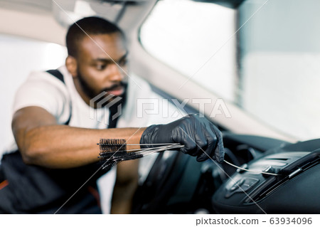 Young handsome concentrated African American man using cleaning brush and removing dust from car air conditioning vent grill. Car detailing or valeting concept. Focus on hand Young handsome concentrated African American man using cleaning brush and removing dust from car air conditioning vent grill. Car detailing or valeting concept. Focus on hand 63934096