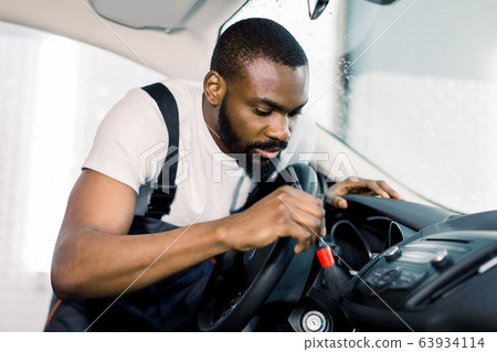 Young African male car wash detailing worker using cleaning brush and removing dust from car control panel. Car detailing or valeting concept. Young African male car wash detailing worker using cleaning brush and removing dust from car control panel. Car detailing or valeting concept. 63934114