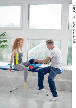 Female patient sitting on couch with knee bolster, male physiotherapist examining her knee Female patient sitting on couch with knee bolster, male physiotherapist examining her knee 63945899