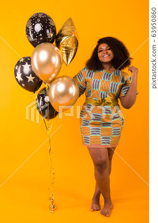 Holidays, birthday party and fun concept - Portrait of smiling young African-American young woman looking sweet on yellow background holding balloons. 63950860