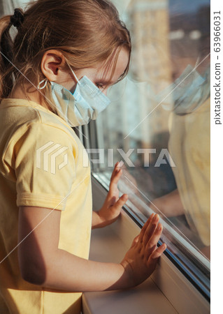 A little girl in a medical mask looks out the window. Coronavirus quarantine. A little girl in a medical mask looks out the window. Coronavirus quarantine. 63966031