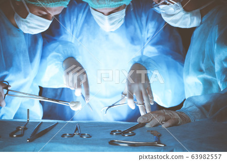 Group of surgeons wearing safety masks performing operation. Close-up of surgery tools. Medicine concept Group of surgeons wearing safety masks performing operation. Close-up of surgery tools. Medicine concept 63982557