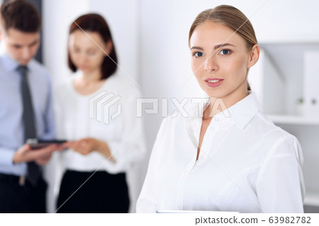 Beautiful blonde businesswoman standing straight in a brightly lit office at the background of colleagues or partners. Business headshot concept Beautiful blonde businesswoman standing straight in a brightly lit office at the background of colleagues or partners. Business headshot concept 63982782