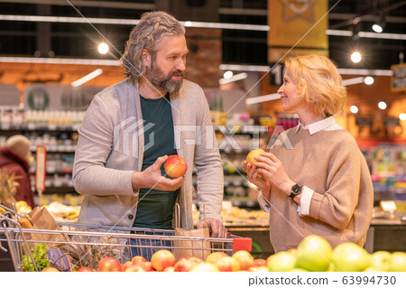 Mature couple with shopping cart discussing sorts of apples in supermarket Mature couple with shopping cart discussing sorts of apples in supermarket 63994730