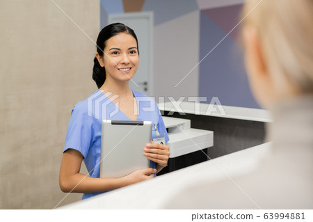 Cheerful young assistant or receptionist in blue uniform looking at patient 63994881