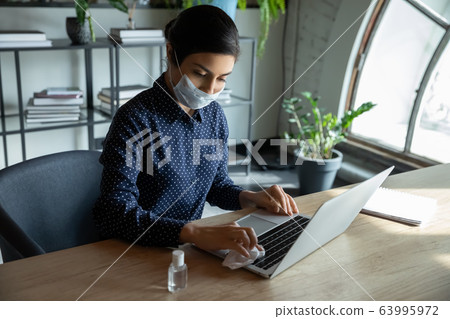 Young businesswoman in facial mask sanitizing laptop keyboard. Young businesswoman in facial mask sanitizing laptop keyboard. 63995972