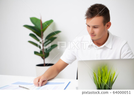 Handsome young worker working in a company office with documents and a laptop 64000051