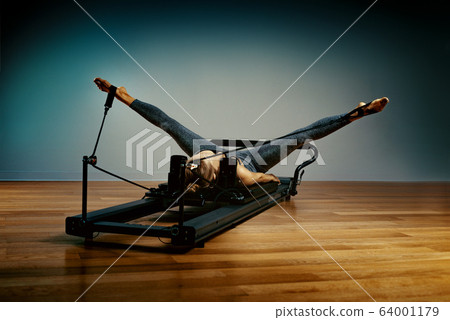 Young girl doing pilates exercises with a reformer bed. Beautiful slim fitness trainer on reformer gray background, low key, art light. Fitness concept Young girl doing pilates exercises with a reformer bed. Beautiful slim fitness trainer on reformer gray background, low key, art light. Fitness concept 64001179