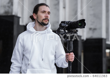 Young Professional videographer holding professional camera on 3-axis gimbal stabilizer. Pro equipment helps to make high quality video without shaking. Cameraman wearing white hoodie making a videos. 64011363
