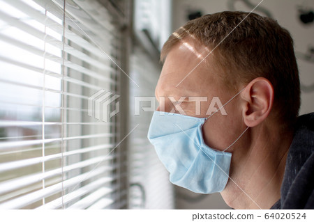 Man in a medical face mask looks out the window through the blinds. Quarantine during an epidemic. 64020524