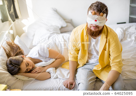 sleepy man in mask sit on bed near sleeping wife 64025095