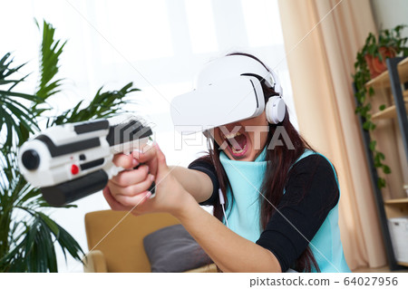 Woman with virtual reality headset and blaster is playing game. 64027956