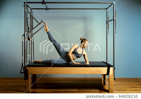Young girl doing pilates exercises with a reformer bed. Beautiful slim fitness trainer on reformer gray background, low key, art light. Fitness concept 64028210