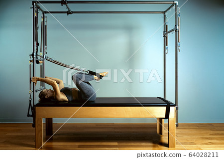 Young girl doing pilates exercises with a reformer bed. Beautiful slim fitness trainer on reformer gray background, low key, art light. Fitness concept 64028211
