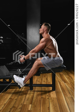 Side view of a strong young man with athletic body doing exercises on fitness equipment in a dark gym. Copy space, advertising banner, fitness motivation, healthy body healthy mind Side view of a strong young man with athletic body doing exercises on fitness equipment in a dark gym. Copy space, advertising banner, fitness motivation, healthy body healthy mind 64028227