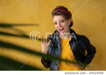 Let's rock concept. Portrait of excited beautiful woman with dark pink hair in casual yellow dress standing, looking at camera, showing rock and roll gesture. 64029496