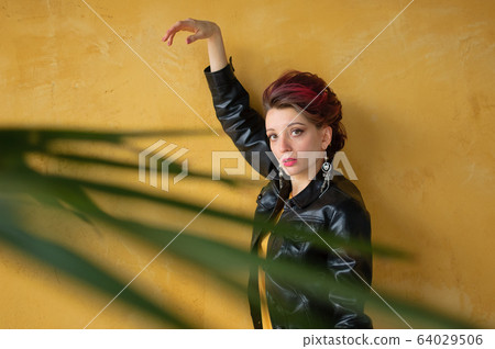 Glamorous lady in punk rock style party clothes with dark pink hair in casual dress, black leather jacket, and long earrings on yellow background 64029506