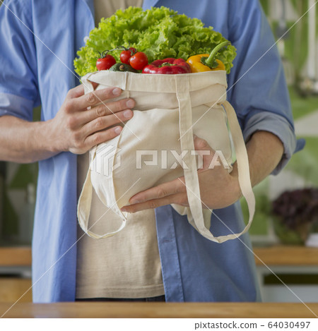 Man holding big cotton reusable bag full of vegetables in the kitchen Man holding big cotton reusable bag full of vegetables in the kitchen 64030497
