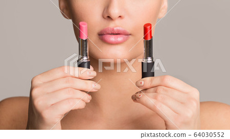 Difficult choice. Girl holding two lipsticks, gray background Difficult choice. Girl holding two lipsticks, gray background 64030552