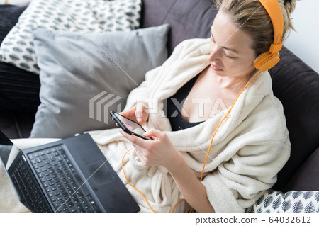 Social distancing. Stay at home. Woman in bathrobe being comfortable at her home sofa, using social media apps on phone for video chatting and stying connected with her loved ones. 64032612