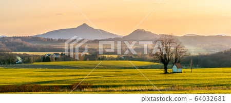 Hilly landscape with small chapel under the tree illuminated by evening sunset. Green grass fields and hills on the horizont. Vivid spring rural countryside. Ralsko Mountain, Czech Republic Hilly landscape with small chapel under the tree illuminated by evening sunset. Green grass fields and hills on the horizont. Vivid spring rural countryside. Ralsko Mountain, Czech Republic 64032681