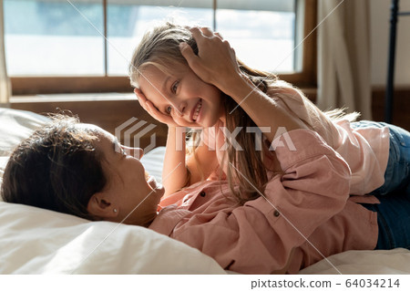Happy mother lying on bed, stroking head of little daughter. 64034214