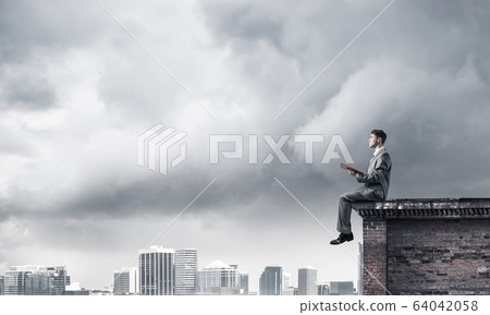 Man on roof edge reading book and cityscape at background 64042058