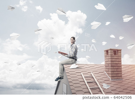 Man on brick roof reading book and paper planes flying in air Man on brick roof reading book and paper planes flying in air 64042506