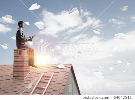 Man on brick roof reading book and paper planes flying in air Man on brick roof reading book and paper planes flying in air 64042511