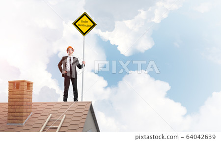 Young businessman on house brick roof holding yellow signboard. Mixed media Young businessman on house brick roof holding yellow signboard. Mixed media 64042639