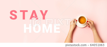 Stay home stamp inscription. Person hands holding a hot cup of tea on a pink background, top view, flat lay. Stay home stamp inscription. Person hands holding a hot cup of tea on a pink background, top view, flat lay. 64051063