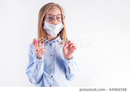 Small pretty girl in a medical mask is holding antiseptic bottle in her hands against light grey background. 64068566