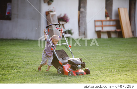 Down syndrome child with lawn mower walking outdoors in garden. 64073299