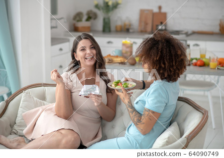 Smiling woman eating chocolate, sad girlfriend with plate of salad. 64090749