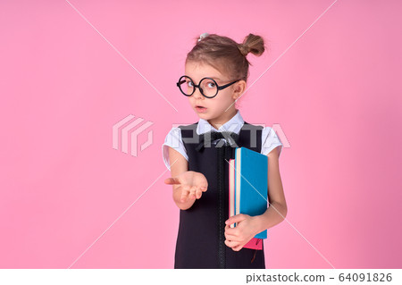 primary school girl in uniform, round glasses without lenses holds a notebook in her hands and lifts her hand to the side with the emotion of a question on her face, posing on a pink background 64091826