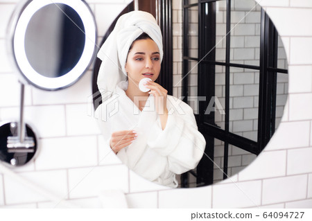 Cute woman in white coat and towel on her head makes herself a makeup Cute woman in white coat and towel on her head makes herself a makeup 64094727