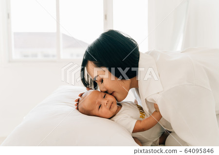 Caring young Asian mum in white wear holding own infant kissing newborn son at home 64095846