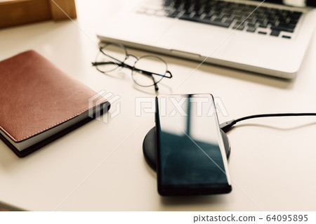 Smart phone being charged on wireless charger alongside with laptop, glasses, and notebook on table. Smart phone being charged on wireless charger alongside with laptop, glasses, and notebook on table. 64095895