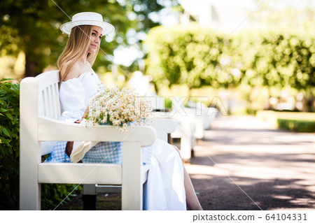 Young woman wearing a hat sitting on a wooden bench and relaxing at the park 64104331