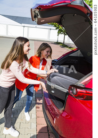 Girls put travel suitcase into big trunk of car 64105983