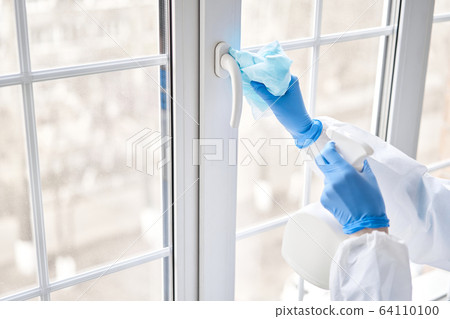 Disinfection, cleaning and washing of handle of the window. A woman in a protective suit and blue gloves sprays a disinfectant solution. Prevention of coronavirus infection. 64110100